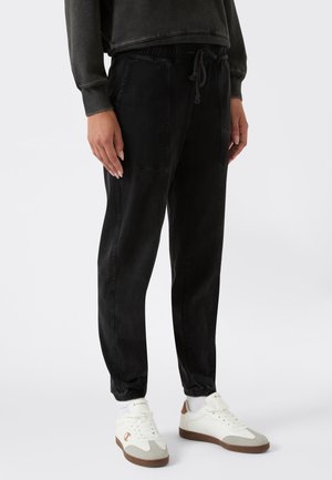 Persona con pantalones jogger negros con puños elásticos y zapatillas blancas con suelas marrones, de pie contra un fondo liso.
