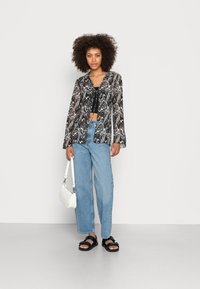 Blusa transparente em preto e branco com padrão ondulado e mangas longas, combinada com jeans de cintura alta azul clara e sandálias pretas, segurando uma bolsa branca.