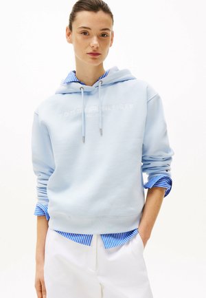 Haut à capuche bleu clair avec le logo "TOMMY HILFIGER". Se distingue par une coupe décontractée, des poignets côtelés et une chemise à rayures bleues et blanches superposée en dessous.