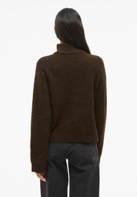Braun gestrickter Pullover mit hohem Kragen, gerippter Textur und lässigen Schultern, kombiniert mit dunklen Hosen. Der Stoff wirkt weich und flauschig.