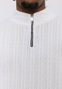 Haut zippé blanc avec un motif texturé en formes de losanges, un col montant et un détail de fermeture éclair noire contrastant.