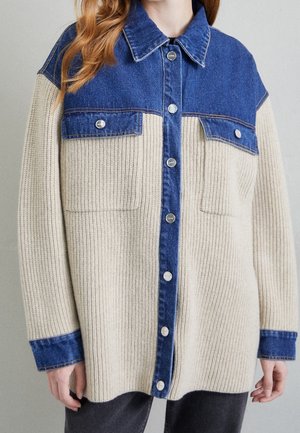 Beżowa prążkowana sweterkowa kurtka zapinana na guziki z niebieskim jeansowym kołnierzem, karczkiem na piersi, mankietami i kieszeniami z patkami, noszona przez osobę z rudymi włosami.