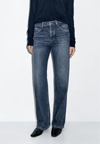 Dark wash denim jeans with a straight leg design, featuring front pockets and subtle stitching details. Paired with black pointed shoes.