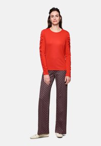 Red long-sleeve top with gathered sides paired with wide-leg patterned trousers in dark brown with diamond shapes in white and red.