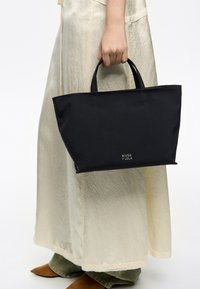 Czarna torba na ramię z solidnymi uchwytami, o prostokątnym kształcie, wykonana z gładkiej tkaniny, z kontrastowymi przeszyciami i logo z przodu.