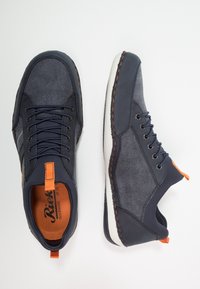 Marinblå avslappnade sneakers med orange dragflikar, detaljerad sömnad och vita sulor, visade från ovan och i profil mot en vit bakgrund.