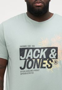 Light green short-sleeve T-shirt with a round neckline, featuring a black and yellow palm graphic, and 'JACK & JONES' branding in bold font.