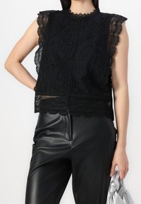 Haut sans manches en dentelle noire avec des bords festonnés, motifs floraux complexes et un col montant, associé à un pantalon en simili cuir noir.