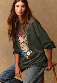 Olivno zelen pulover prevelikega kroja z grafiko cvetja Frida Kahlo, z belim besedilom in pisanimi cvetlicami, ki okrašujejo dizajn.