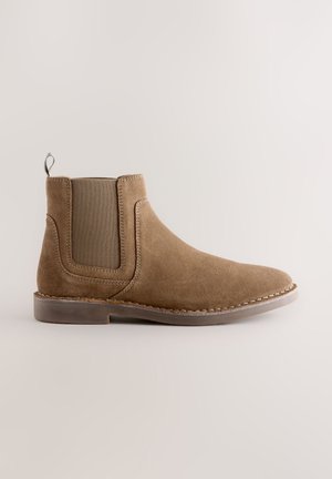 Brun ruskind Chelsea ankelstøvle med elastisk sidepanel, trækstrop ved hælen og lav stablet gummisål på neutral baggrund.