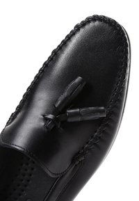 Mocassin en cuir noir avec garniture tressée et détail pompon sur le dessus, montrant une texture lisse et des bords cousus.