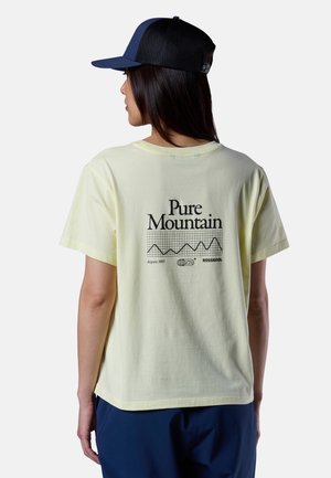 Donna che indossa una T-shirt giallo chiaro con la scritta "Pure Mountain" e grafica di montagna, e un cappellino nero in rete, di spalle su uno sfondo bianco semplice.