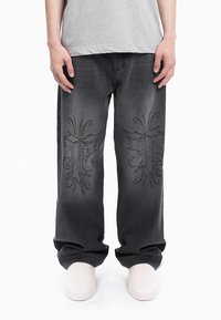 Calças de ganga pretas largas com designs bordados decorativos nas pernas exteriores. Acabadas com uma lavagem desbotada e estilo tradicional de cinco bolsos.