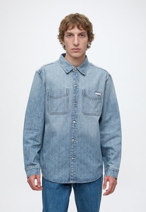 Junger Mann mit lockigem hellbraunem Haar, der ein hellblaues Jeanshemd mit Knöpfen und blaue Jeans trägt, vor einem schlichten weißen Hintergrund stehend.