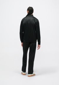 Žmogus su pintinėmis plaukais, vilkintis juodą treniruoklių kostiumą ir baltus sportbačius, stovi nugara pasukęs priešais vienspalvę baltą fono.