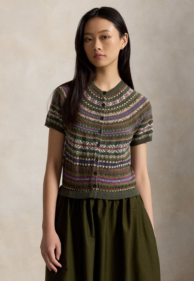 Jeune femme aux longs cheveux foncés portant un cardigan vert à motifs à manches courtes boutonné et une jupe vert foncé sur un fond neutre.