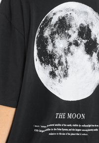 Sort bomulds T-shirt med et stort hvidt grafisk design af månen, med tekst nedenunder der definerer "Månen" og dens egenskaber.