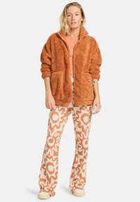 Veste en polaire brun avec deux poches avant, associée à un pantalon évasé crème et orange à motif floral. Texture douce.