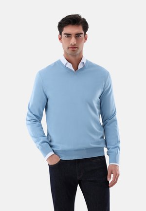 Hellblaues V-Ausschnitt-Pullover aus glattem Strickstoff, mit gerippten Bündchen und Saum; kombiniert mit einem weißen Hemd und dunklen Jeans.