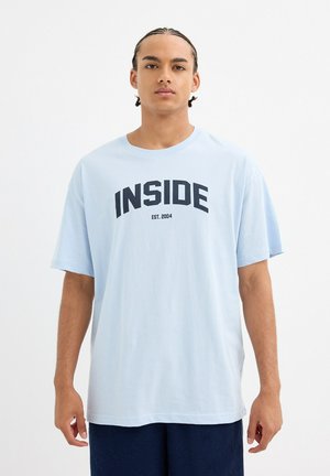 Camiseta de algodón de color azul claro con mangas cortas, que presenta una gran impresión en negro con el texto "INSIDE" y "EST. 2004" centrado debajo.
