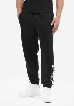 Mann trägt schwarze Jogginghosen mit weißem "London"-Schriftzug am Unterschenkel, ein schwarzes Hemd, weiße Socken und weiße Sneaker, steht lässig.