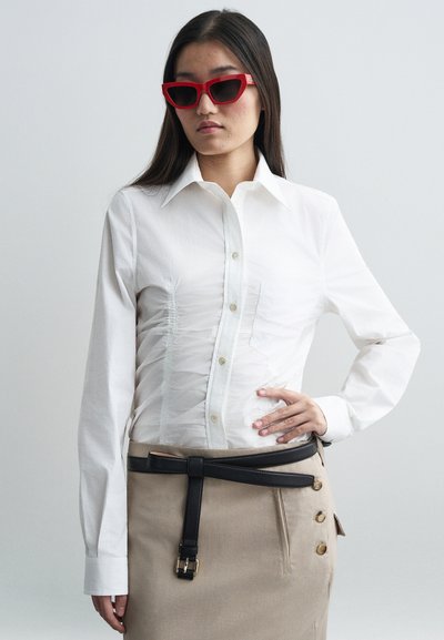 Camisa blanca con cuello tipo button-down, con detalles de fruncido texturizados; combinada con una falda beige y un cinturón negro con hebillas doradas. Gafas de sol rojas.