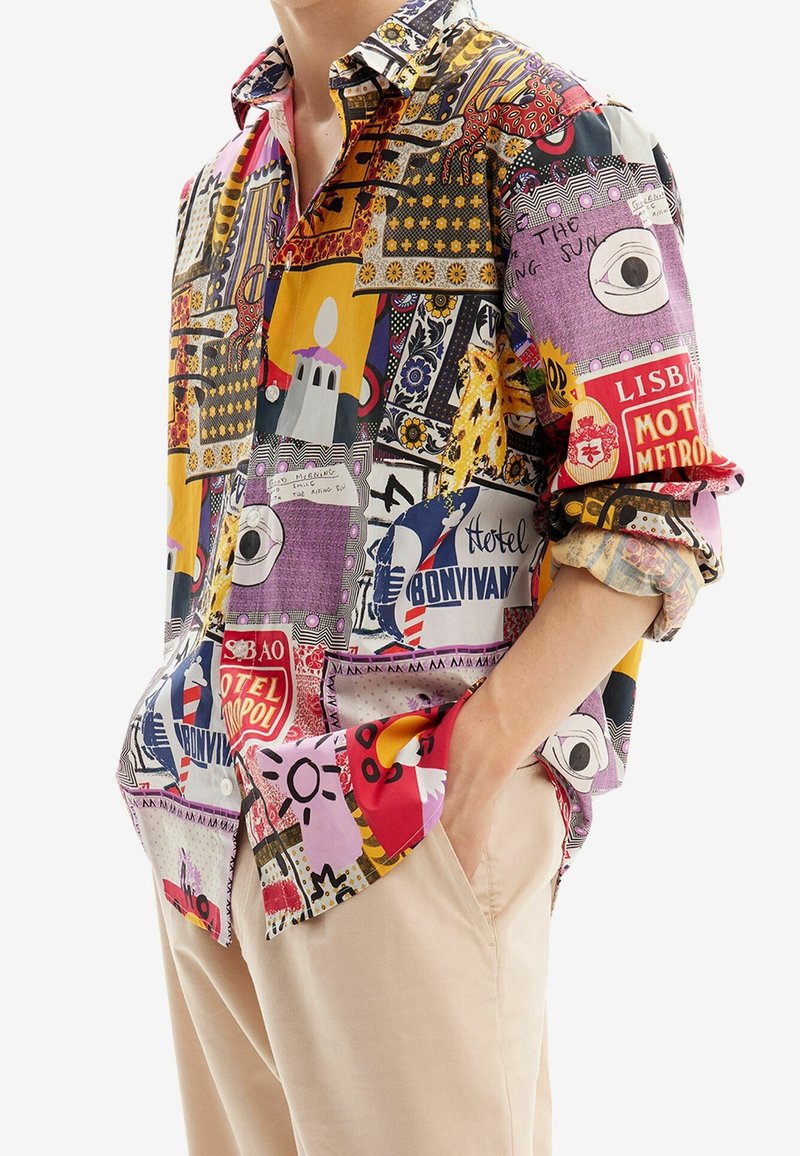 Homme portant une chemise colorée à motif patchwork boutonnée avec les manches retroussées et un pantalon beige, debout avec une main dans la poche.