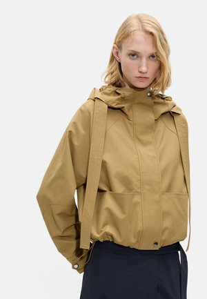 Młoda kobieta o jasnych włosach, ubrana w jasnobrązową kurtkę z kapturem i dużymi paskami oraz ciemne spodnie, stoi twarzą do przodu z neutralnym wyrazem twarzy.