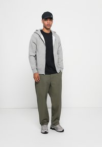 Grijze zip-up hoodie over een zwart T-shirt, gecombineerd met loose olivegroene broek en grijze sneakers, afgemaakt met een zwarte pet.