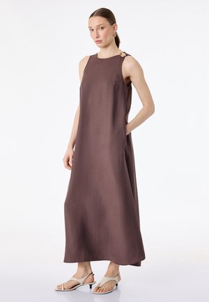 Kobieta w brązowej, bezrękawowej sukience maxi z klamrą na ramieniu, beżowych sandałkach na obcasie i złotych kolczykach kołach, stojąca na białym tle.