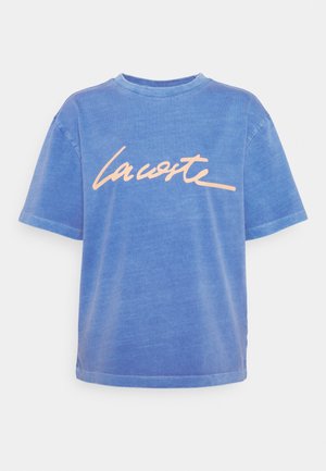 T-shirt bleu à manches courtes avec une texture délavée et un grand logo cursif « Lacoste » de couleur pêche claire sur la poitrine.