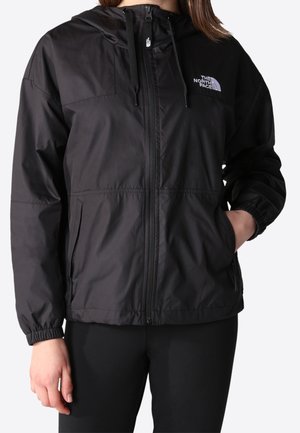 Veste coupe-vent noire zippée avec capuche et logo sur la poitrine portée par une personne avec une main dans la poche et l'autre détendue sur le côté.