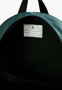 El interior de la mochila en tonos teal presenta un forro negro con una etiqueta blanca para nombre y datos de contacto, contorneada con costuras negras. Bolsillo de malla en la parte inferior.