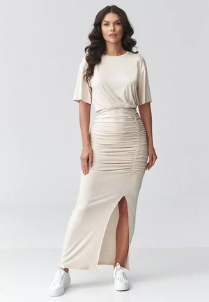 Kobieta z długimi, ciemnymi kręconymi włosami, ubrana w beżową marszczoną sukienkę z przednim rozcięciem i białe trampki, stojąca na tle jednolitego, jasnego tła.