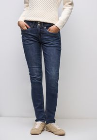 Mörkblå denimjeans med smal passform, utrustade med fem fickor, knapplåsning och dragkedja. Kombineras med beige sneakers och en krämfärgad kabelstickad tröja.