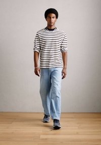 Jovem andando para a frente, vestindo uma camisa de manga longa branca e preta com riscas, jeans azul claro e ténis cinzentos, sobre um chão de madeira.