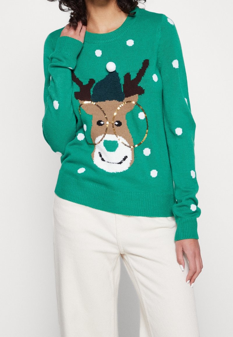 Pull vert tricoté avec un motif de renne orné de pois blancs. Le renne porte un chapeau bleu et des anneaux dorés autour de son nez.