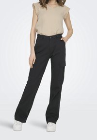 Femme portant un haut sans manches beige et un pantalon cargo noir avec des poches latérales, associé à des baskets blanches.