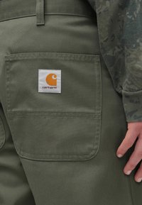 Zielone spodnie z płótna z tylną kieszenią, na której znajduje się biała metka z pomarańczowym logo. Detale przeszyć podkreślają trwałość i design.