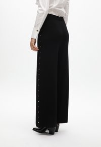 Žena v černých širokých kalhotách s vysokým pasem a zlatými bočními hroty, bílé blůze a černých jehlových botách s špičatou špičkou, stojící na bílém pozadí.