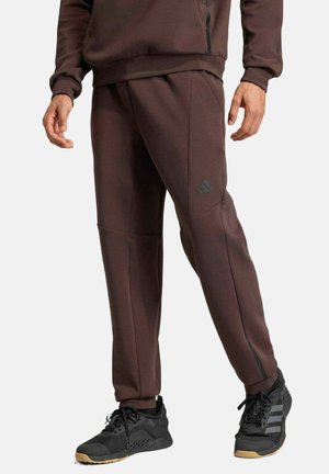 Man draagt bruine Adidas joggingbroek en bijpassende sweatshirt met zwarte Adidas sneakers, staand tegen een effen achtergrond.