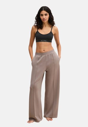 Vrouw met krullend donker haar, gekleed in een zwarte sportbeha en losse taupekleurige broek met wijde pijpen, staand op blote voeten met de handen in de zakken.