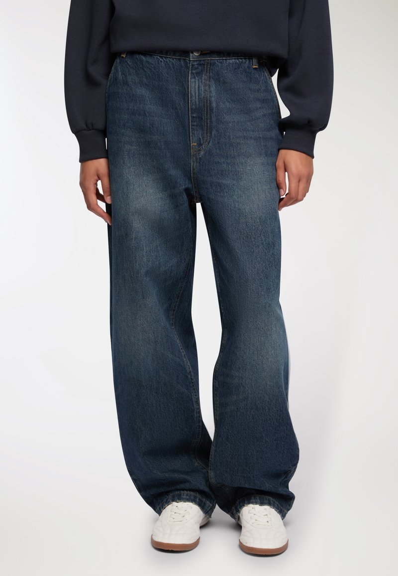 Szerokie, ciemnoniebieskie dżinsy wykonane z denimu, z wysokim stanem, przednimi kieszeniami i delikatnym przetarciem, w połączeniu z białymi sneakersami.