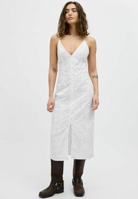 Vestido maxi blanco texturizado con escote en V, tirantes finos y botones en el frente; complementado con botas largas de cuero marrón con hebillas.