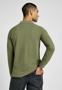 Olivgrön långärmad polo med krage, med en diskret vit kant, tillverkad av ett slätt tyg, kombinerad med mörka jeans.