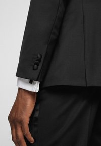 Giacca nera su misura con un taglio elegante, caratterizzata da tre bottoni neri sul polsino e una camicia bianca che sbuca sotto.