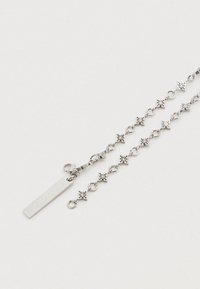 Ασημένιο κολιέ με αλυσίδα που έχει συνδέσμους σε σχήμα αστεριού, επίπεδη ταμπέλα με χάραξη και κούμπωμα με κρίκο τύπου αστακός. Ομοιόμορφη μεταλλική υφή σε όλο το μήκος.