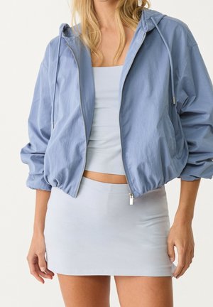 Woman wearing light blue cropped zip-up hoodie, matching tube top, and mini skirt with long blonde hair visible.