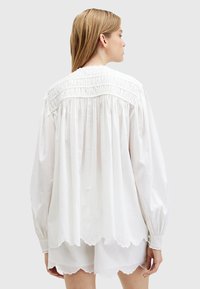 Blusa bianca in cotone con yoke arricciato, maniche lunghe a palloncino e orlo a zigzag. Tessuto morbido con pieghe sul retro.