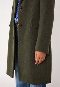 Cappotto in lana verde oliva con finitura texturizzata, dotato di quattro bottoni, revers e tasche laterali, indossato sopra un jeans.